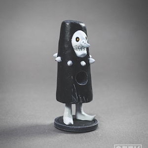 Op de afbeelding: Zwart-witte handgeschilderde figuur van een skelet met een grote neus en een zwarte robe. De figuur heeft een rond gat in zijn borst en staat op een zwarte basis.