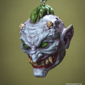 May include: A white and purple ceramic ornament depicting a grotesque, grinning demon with green eyes, a green mohawk, and sharp teeth. The ornament is hanging from a string.