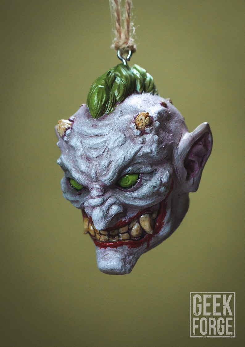 May include: A white and purple ceramic ornament depicting a grotesque, grinning demon with green eyes, a green mohawk, and sharp teeth. The ornament is hanging from a string.