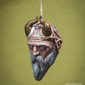 May include: A hanging ornament depicting a detailed, sculpted head of a bearded man with horns and runes on his forehead. The ornament is made of a light brown material and has a rope attached to the top.