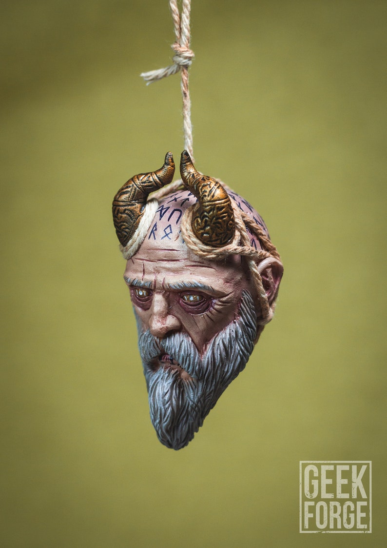 May include: A hanging ornament depicting a detailed, sculpted head of a bearded man with horns and runes on his forehead. The ornament is made of a light brown material and has a rope attached to the top.