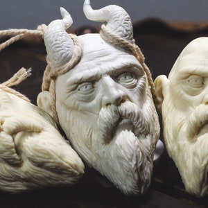 May include: Three white carved wooden sculptures of bearded men's faces. Two of the sculptures have horns on their heads. The sculptures are on a dark wooden surface.