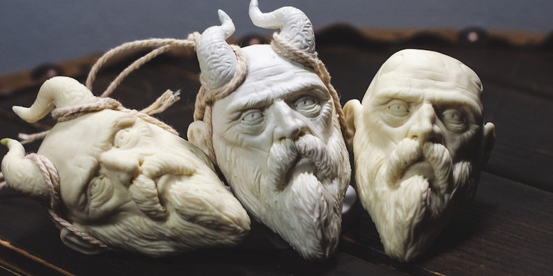 May include: Three white carved wooden sculptures of bearded men's faces. Two of the sculptures have horns on their heads. The sculptures are on a dark wooden surface.