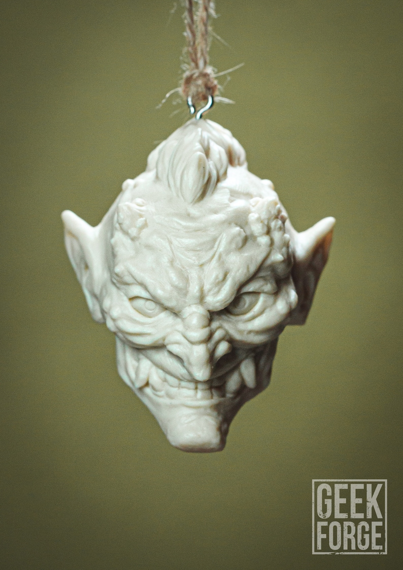 May include: A white ceramic ornament shaped like a goblin's head with a menacing expression. The ornament has a loop at the top for hanging.