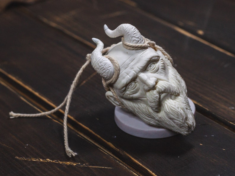 May include: White ceramic sculpture of a bearded man's head with horns, on a wooden surface.