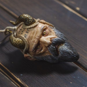 May include: A sculpted head with a beard and horns, the face is painted with runes and has a weathered look. The head is lying on a dark wood surface.