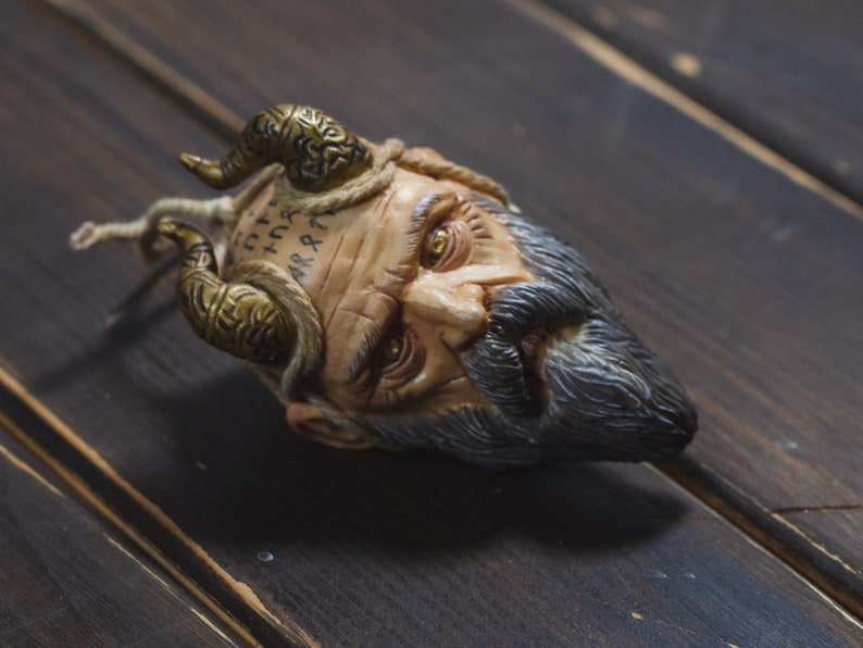 May include: A sculpted head with a beard and horns, the face is painted with runes and has a weathered look. The head is lying on a dark wood surface.