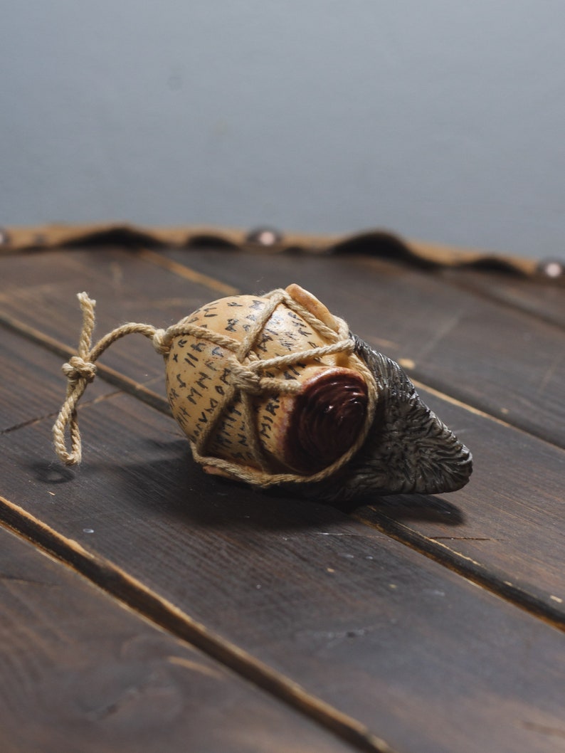 May include: A small, round, brown and white object wrapped in twine with runes inscribed on the surface. The object is tied with a brown rope and has a dark brown, textured, animal-like tail.