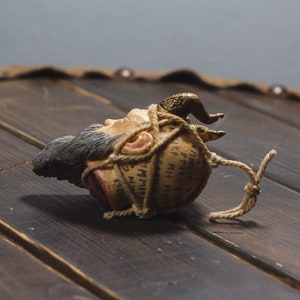 May include: A small, brown, ceramic head with a beard and horns is wrapped in twine and has writing on it. The head is lying on a wooden surface.