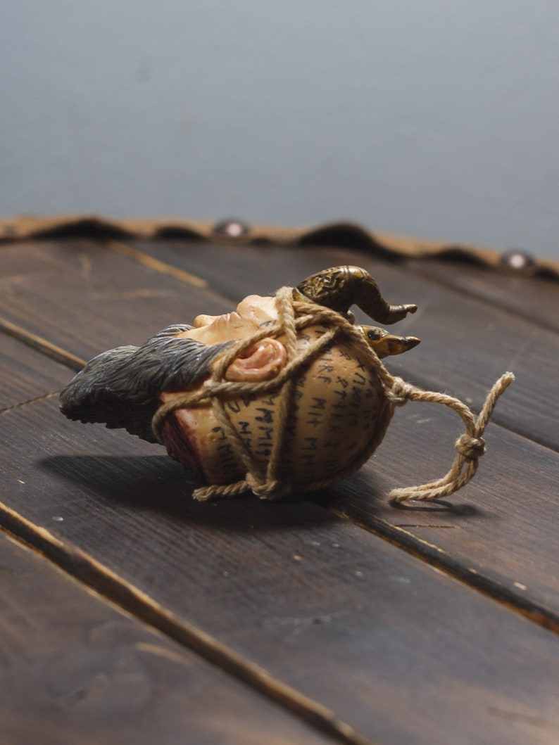 May include: A small, brown, ceramic head with a beard and horns is wrapped in twine and has writing on it. The head is lying on a wooden surface.