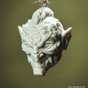 May include: A white ceramic ornament shaped like a goblin's head with a menacing expression. The ornament has a textured surface and is hanging from a brown twine string.