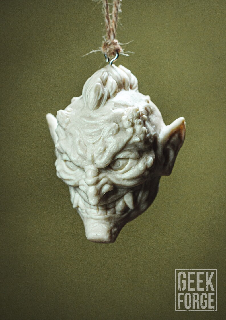 May include: A white ceramic ornament shaped like a goblin's head with a menacing expression. The ornament has a textured surface and is hanging from a brown twine string.