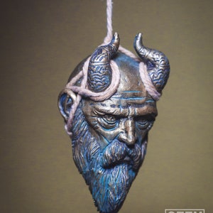 May include: A detailed, sculpted, hanging ornament of a bearded man's head with horns. The head is painted in shades of blue and gold, with a rope tied around the horns. The ornament is hanging from a rope.