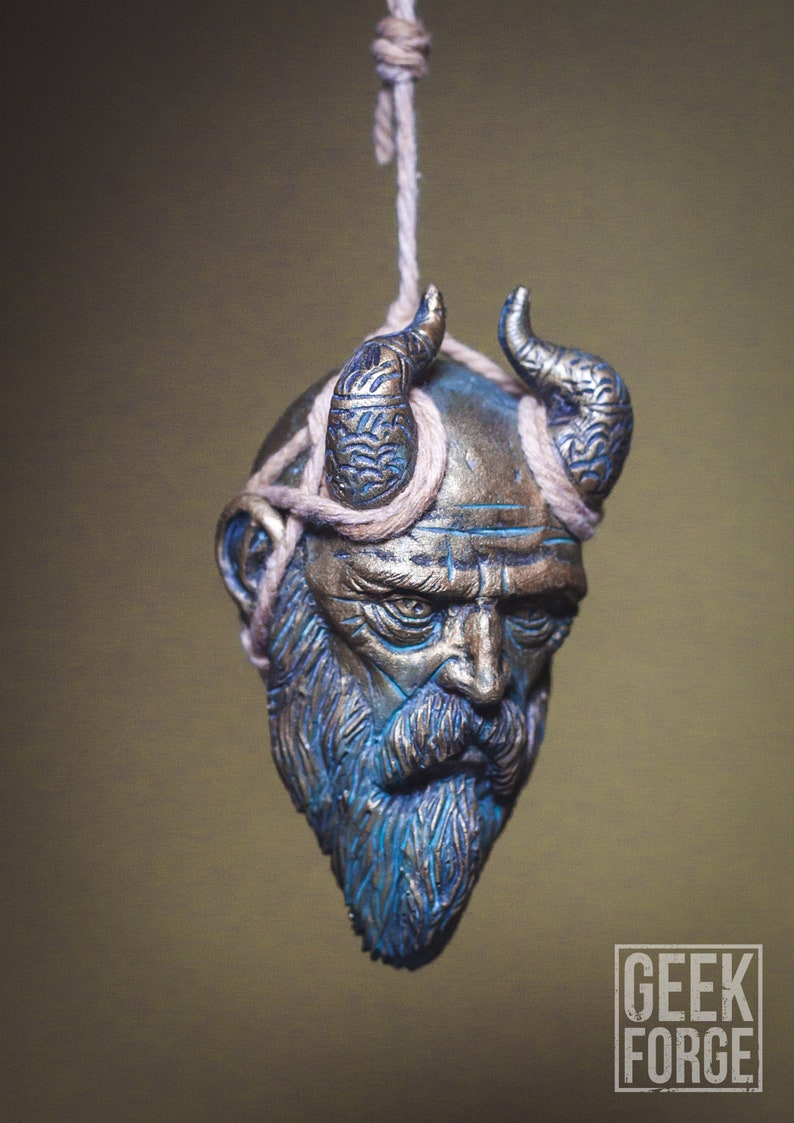 May include: A detailed, sculpted, hanging ornament of a bearded man's head with horns. The head is painted in shades of blue and gold, with a rope tied around the horns. The ornament is hanging from a rope.