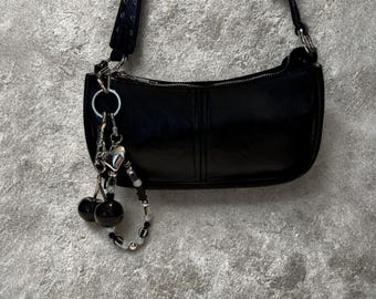 May include: Black shoulder bag with a curved shape and a silver zipper. The bag has a decorative charm with a heart, beads, and cherry-shaped accents. The strap is black and adjustable.