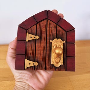 May include: A miniature wooden door with a red frame and a brown door. It features gold-coloured hinges and a doorknob. The design mimics a small house door.