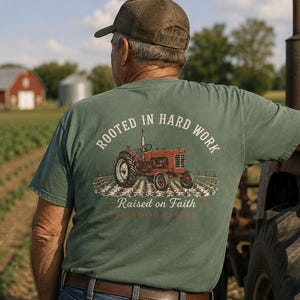 Può includere: Una t-shirt a maniche corte color verde salvia con una grafica sul retro raffigurante un trattore rosso in un campo. Il testo sopra il trattore recita Rooted in Hard Work e il testo sottostante recita Raised on Faith in carattere bianco e rosso.