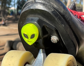 Pode incluir: Close-up de um patins de rodas preto com um adesivo de rosto de alienígena verde no freio. O patins tem uma roda branca com a palavra "Fame" impressa nela.