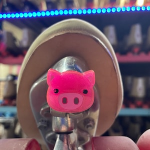 May include: A pink pig-shaped roller skate stopper. The stopper is attached to a silver metal skate truck with a red wheel.