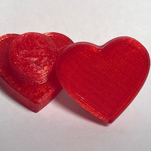 Puede incluir: Dos cajas rojas en forma de corazón, una apilada sobre la otra. La caja superior es ligeramente más pequeña que la caja inferior.