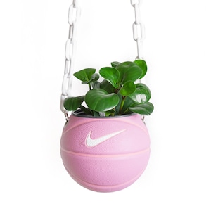 Pode incluir: Um vaso de planta de basquete rosa com um logotipo Nike branco em forma de swoosh pendurado em uma corrente branca. Uma planta verde está crescendo dentro do vaso.