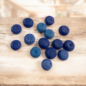 May include: A collection of round, dark blue wax melts resembling blueberries, scattered on a light brown wooden surface. The wax melts vary slightly in shade, with some being a lighter blue. The background is blurred, suggesting a natural setting.