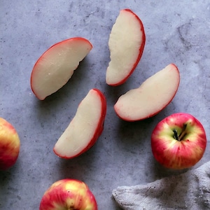 May include: Overhead shot of fresh apples and apple slices on a gray surface. The apples are red and yellow, with some cut into wedges. The apple slices have a red skin and white flesh. A gray cloth is in the bottom right corner.