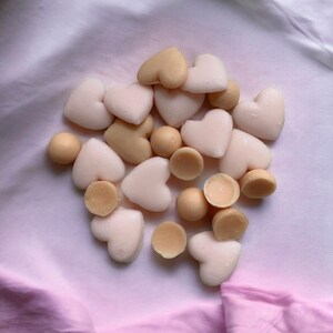 May include: A collection of heart-shaped and round, light-colored wax melts. The melts are in shades of cream and beige, scattered on a pink and white fabric background. Some melts are whole, while others are cut in half.