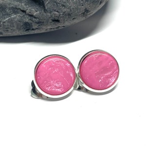 Könnte beinhalten: Ein Paar Clip-Ohrringe mit einem hellrosa, kreisförmigen Design, das in einen silberfarbenen Metallrahmen gefasst ist. Die Ohrringe haben eine glatte, glänzende Oberfläche und ein schlichtes, elegantes Design. Der Clip-Mechanismus ist sichtbar.