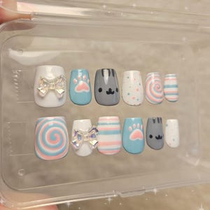 Puede incluir: Un juego de uñas postizas en una caja de plástico transparente. Las uñas presentan varios diseños, incluyendo lazos, huellas de patas, caras de gato y patrones de remolino en colores pastel. Las uñas son de forma cuadrada.