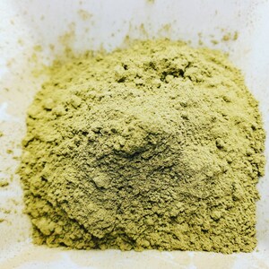 May include: A mound of fine, dry, olive-green powder with a matte texture. The substance is granular and appears uniform in color, forming a soft, rounded heap that occupies the center of the frame with visible small clumps throughout the pile.