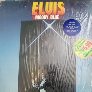 Puede incluir: Un &aacute;lbum de vinilo "Moody Blue" de Elvis Presley. La portada del &aacute;lbum presenta el t&iacute;tulo en naranja y el nombre del artista en amarillo. Una pegatina en el &aacute;lbum indica que contiene los &eacute;xitos "Moody Blue" y "Way Down".
