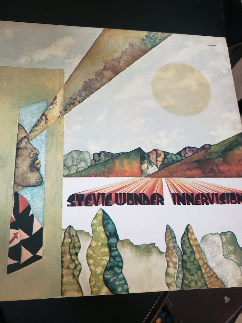 Puede incluir: Una portada de &aacute;lbum de vinilo vintage de Stevie Wonder, "Innervisions". La portada es un dise&ntilde;o abstracto colorido con la cara de un hombre asom&aacute;ndose desde el lado izquierdo de la imagen. El fondo presenta un sol amarillo brillante, un cielo azul y un paisaje con &aacute;rboles y colinas.