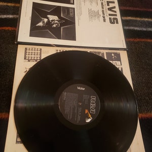 May include: A black vinyl record with a white label that says "RCA Victor" and "Elvis As Recorded At Madison Square Garden". The record is on a white cardboard sleeve with a black and white photo of Elvis Presley on the front.