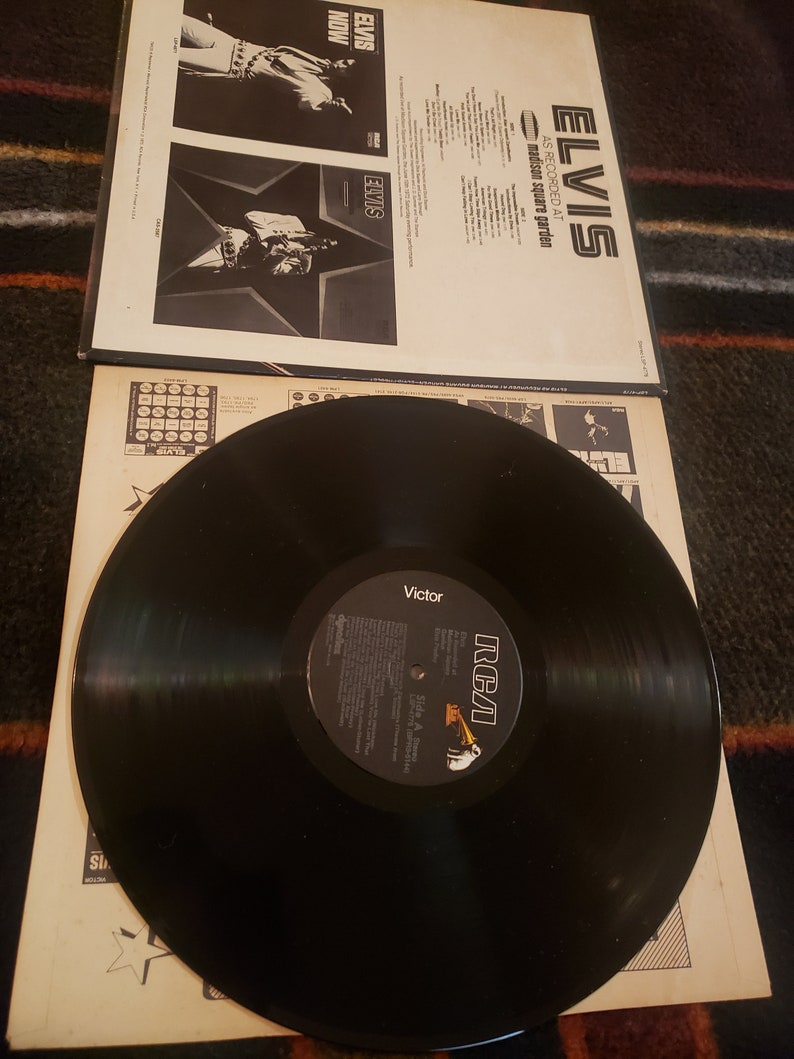 May include: A black vinyl record with a white label that says "RCA Victor" and "Elvis As Recorded At Madison Square Garden". The record is on a white cardboard sleeve with a black and white photo of Elvis Presley on the front.