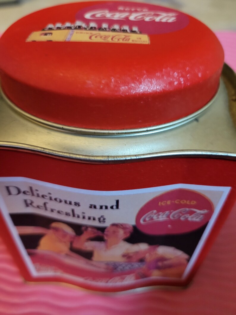 Puede incluir: Lata roja de Coca-Cola con una imagen vintage de dos personas disfrutando de Coca-Cola con hielo. La lata tiene el texto "Delicious and Refreshing" y "Ice-Cold Coca-Cola".