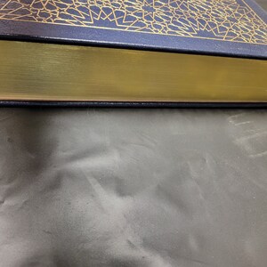 Puede incluir: Un libro de tapa dura de color azul marino con detalles dorados y un lomo dorado. El libro est&aacute; cerrado y sobre una superficie gris.