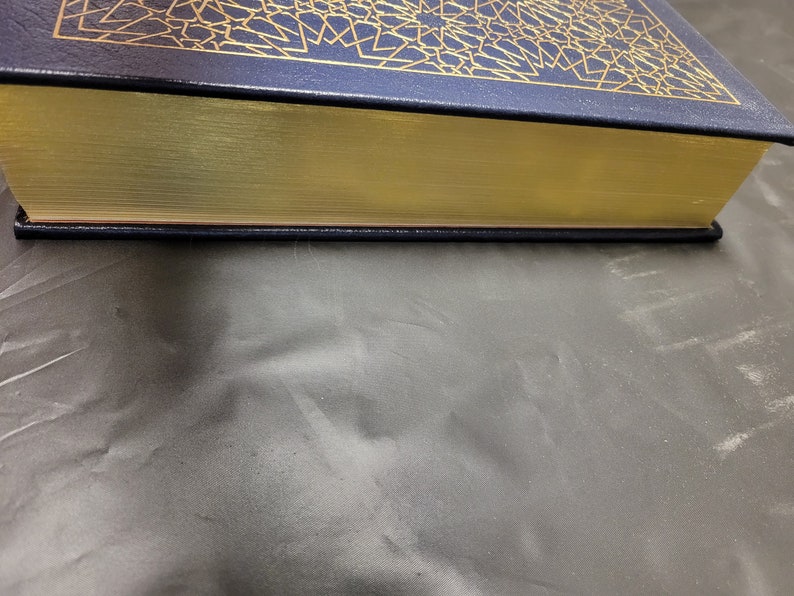 Puede incluir: Un libro de tapa dura de color azul marino con detalles dorados y un lomo dorado. El libro est&aacute; cerrado y sobre una superficie gris.