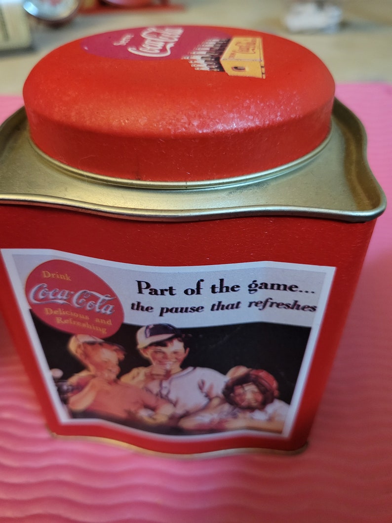 Puede incluir: Lata roja de Coca-Cola con tapa plateada. La lata tiene un logotipo vintage de Coca-Cola y el texto "Drink Coca-Cola Delicious and Refreshing Part of the game... the pause that refreshes". La lata presenta una imagen vintage de tres personas disfrutando de una Coca-Cola.