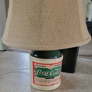 May include: A green and white ceramic Coca-Cola lamp base with a burlap lampshade. The lamp base features the Coca-Cola logo and the text "Delicious, Home Office Atlanta, Refreshing".