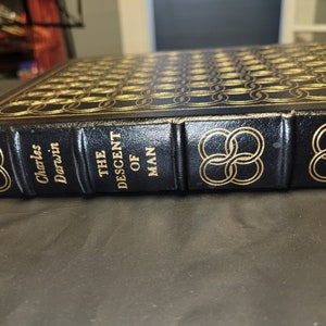 May include: A black leather-bound book with gold lettering and a gold design on the cover. The title of the book is "The Descent of Man" by Charles Darwin.