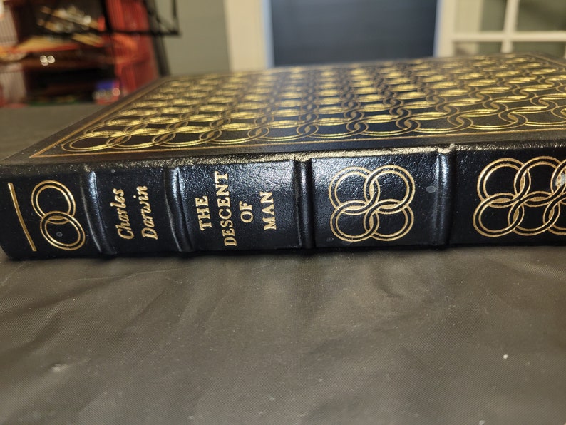 May include: A black leather-bound book with gold lettering and a gold design on the cover. The title of the book is "The Descent of Man" by Charles Darwin.