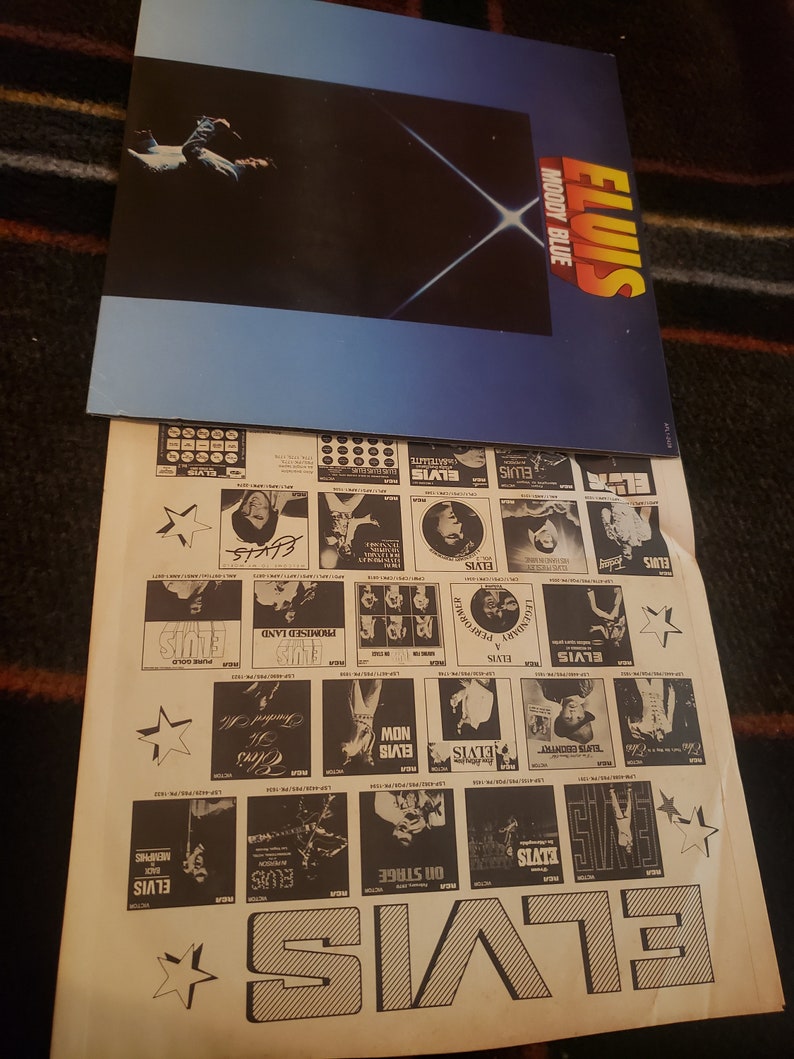 Puede incluir: Una portada del &aacute;lbum "Moody Blue" de Elvis Presley con un fondo azul y negro y un dise&ntilde;o de explosi&oacute;n de estrellas blancas. Debajo del &aacute;lbum hay un p&oacute;ster promocional con varias portadas de &aacute;lbumes de Elvis y el nombre "ELVIS" en letras grandes.