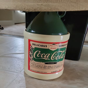 May include: A vintage-style ceramic Coca-Cola jar with a green lid. The jar has a white background with red and green lettering that reads "Delicious Coca-Cola Refreshing Home Office Atlanta" and instructions for dispensing the drink.