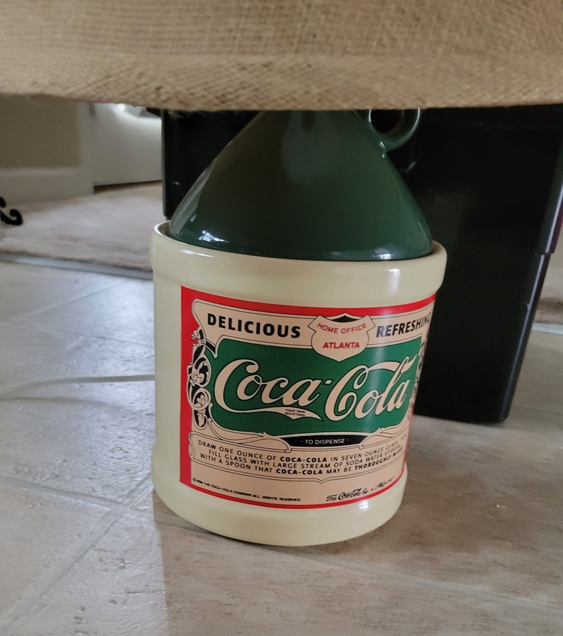 May include: A vintage-style ceramic Coca-Cola jar with a green lid. The jar has a white background with red and green lettering that reads "Delicious Coca-Cola Refreshing Home Office Atlanta" and instructions for dispensing the drink.