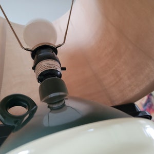 May include: A green ceramic table lamp with a white shade. The lamp has a handle on the side and a light bulb is visible inside the shade.