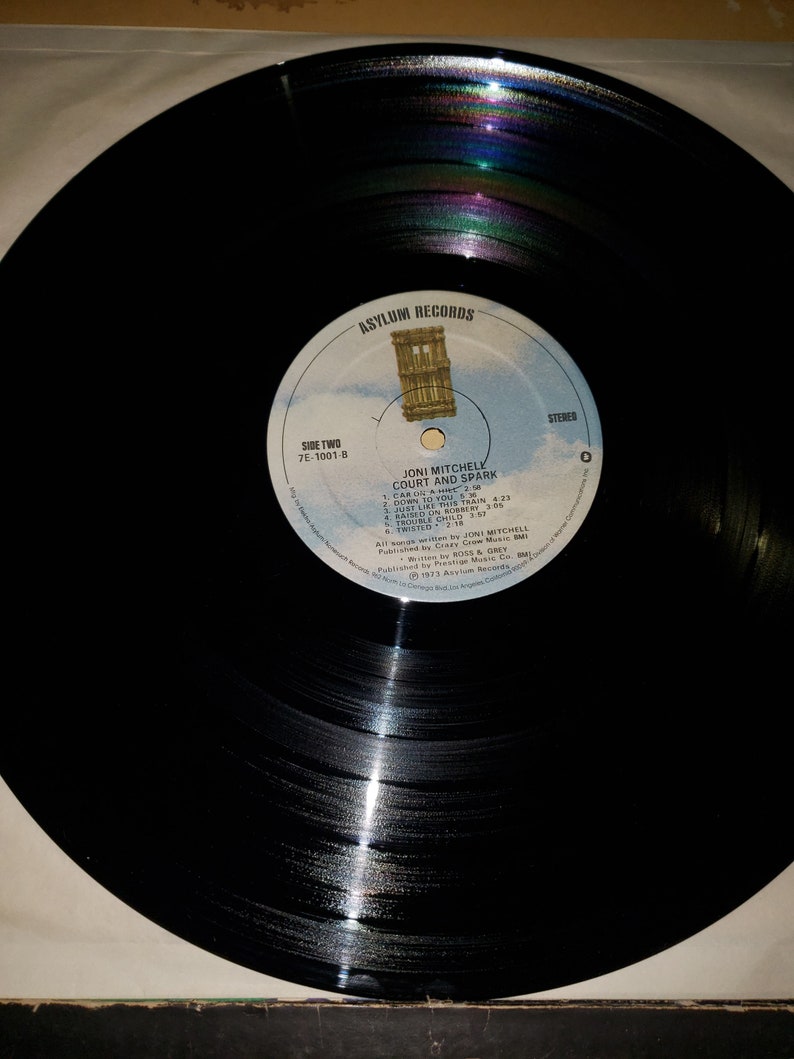 Puede incluir: Disco de vinilo negro con una etiqueta blanca. La etiqueta tiene el texto "Asylum Records", "Side Two", "7E-1001-B", "Joni Mitchell", "Court and Spark", "Car On A Hill", "Down To You", "Just Like This Train", "Raised On Robert", "Trouble Child", "Twisted", "All songs written by Joni Mitchell", "Published by Crazy Crow Music, BMI", "RO55 & GREY", "℗ 1973 Asylum Records", "Manufactured by Asylum Records, 4250  Camino  Real, Los Angeles, California 90065", "Stereo", y "0".