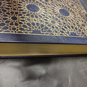 Puede incluir: Un libro de tapa dura de color azul marino con un dise&ntilde;o geom&eacute;trico dorado en la portada. El libro tiene un lomo dorado y un borde dorado.