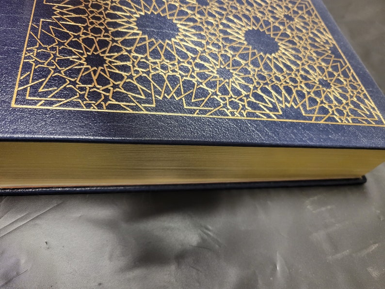 Puede incluir: Un libro de tapa dura de color azul marino con un dise&ntilde;o geom&eacute;trico dorado en la portada. El libro tiene un lomo dorado y un borde dorado.