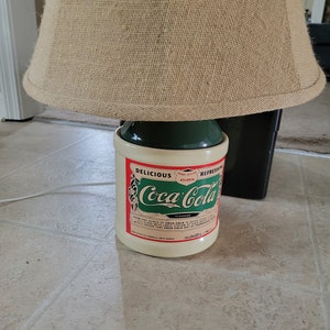 May include: A vintage Coca-Cola lamp base with a green and white ceramic bottle design. The bottle has a red label with the text "Delicious and Refreshing Coca-Cola" and "Home Office Atlanta". The lamp has a beige burlap lampshade.